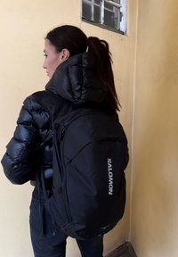 Sac à dos Salomon noir avec une texture lisse et mate ; équipé de sangles ajustables et d'un logo bien visible sur le côté. Porté par une personne portant une veste matelassée.