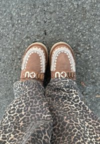 Καφέ σουέτ παπούτσια με διακοσμητική κρεμ ραφή και χρυσό λογότυπο "τώρα", φορεμένα με παντελόνι λεοπάρ, τοποθετημένα σε τραχιά επιφάνεια ασφάλτου.