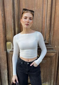 Witte lange mouwen crop top met een aangesloten ontwerp, gecombineerd met hoge zwarte jeans. De achtergrond toont een houten deur.