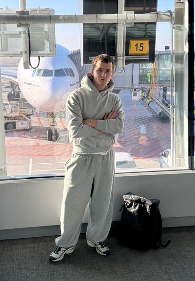 Sudadera gris con capucha y pantalones de chándal a juego, ajuste casual. Mochila negra en el suelo. Ambiente de aeropuerto con un avión Boeing de fondo.