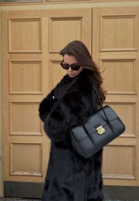 Femme en manteau de fourrure noire et lunettes de soleil, portant un grand sac à main en cuir noir, debout devant une porte en bois à panneaux.