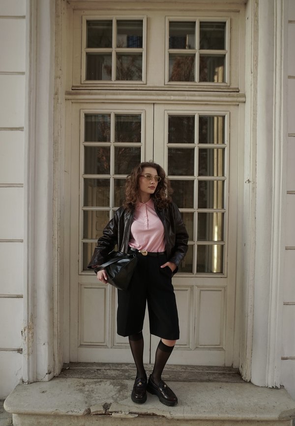 Kvinne med krøllet hår som bruker briller, rosa skjorte, svart skinnjakke, svarte shorts og loafers, står foran en dør med glassruter.