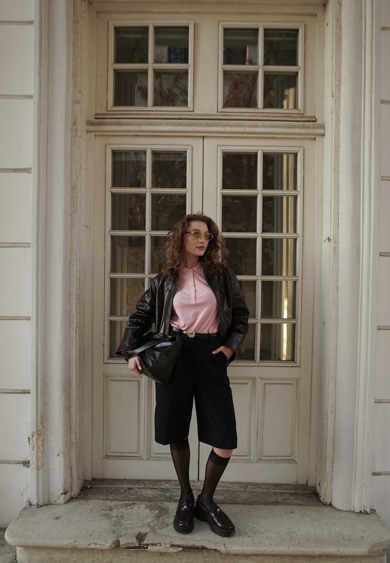 Žena s kučeravými vlasmi, ktorá nosí okuliare, ružovú košeľu, čiernu koženú bundu, čierne šortky a mokasíny, stojí pred sklennými dverami s panelmi.