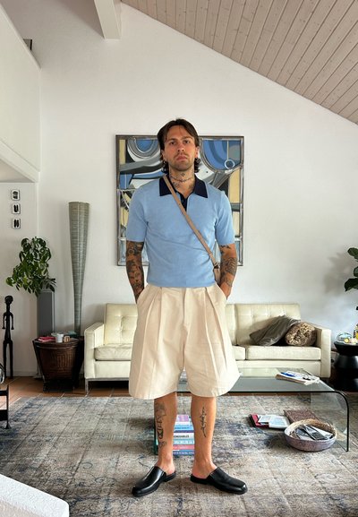 Polo azul claro con collar marino, pantalones cortos beiges con pliegues y zapatos negros sin cordones. De pie en un entorno interior moderno con muebles y decoración.