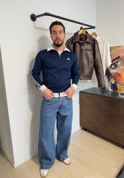 Hombre con cabello rizado y barba, vistiendo chaqueta deportiva azul marino y jeans holgados, de pie junto a una chaqueta de cuero marrón colgada y una camisa blanca.