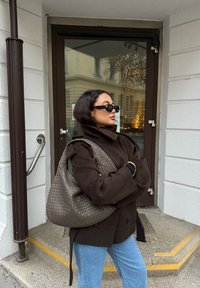 Manteau en laine marron avec col montant, jean bleu clair, lunettes de soleil noires, et sac à main tressé marron texturé. En arrière-plan, une porte en verre.