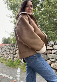 Kvinne iført en brun lammeskinnsjakke og blå jeans som står utendørs nær en steinmur med grønne trær i bakgrunnen.