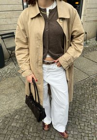 Μπεζ παλτό πάνω από καφέ κάρντιγκαν, λευκά παντελόνια με φαρδιά πόδια και καφέ παπούτσια με μπορντό λεπτομέρειες. Μαύρη τσάντα στο χέρι.