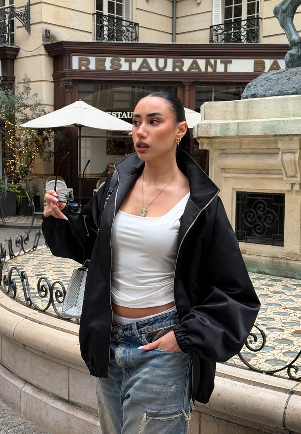 Ung kvinne iført svart jakke og hvit topp holder solbriller nær en restaurantinngang med uteservering.