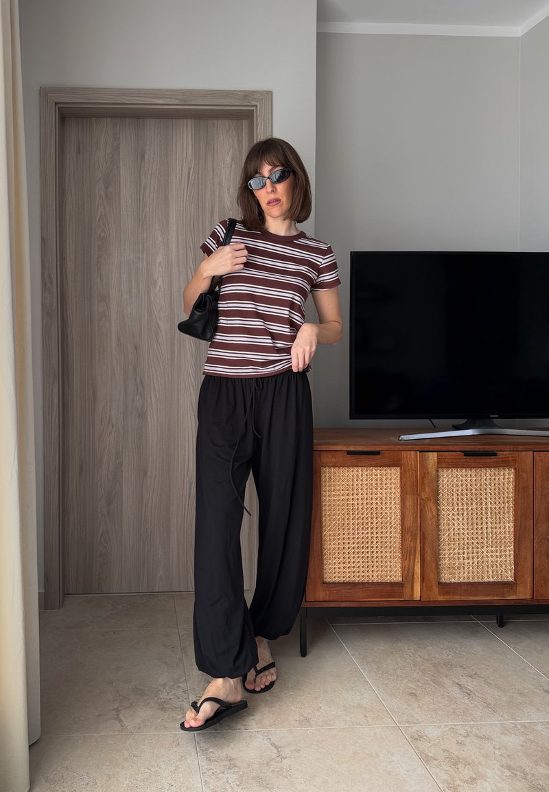 Frau trägt ein gestreiftes T-Shirt, schwarze Hose, Sonnenbrille und Flip-Flops und posiert drinnen neben einem Holzschrank mit einem Flachbildfernseher darauf.