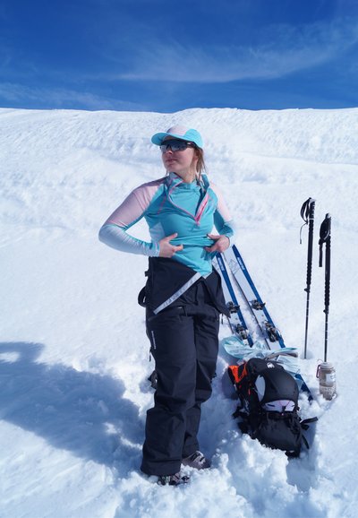 Mujer con equipo de esquí ajustándose la chaqueta en la nieve, con esquís, bastones, mochila y botella de agua a su lado bajo un cielo azul despejado.