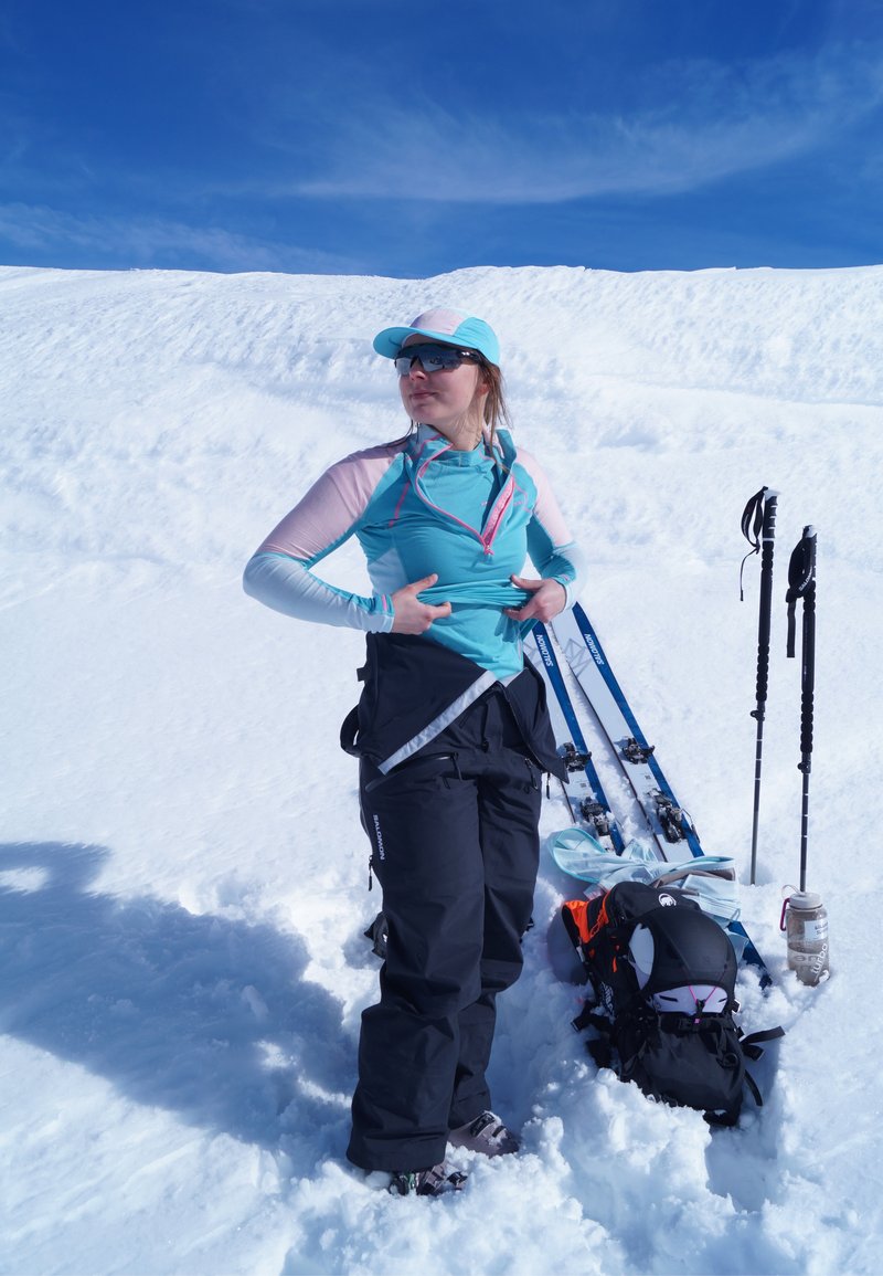Femme en tenue de ski ajustant sa veste dans la neige, avec ses skis, bâtons, sac à dos et bouteille d'eau à côté, sous un ciel bleu clair.