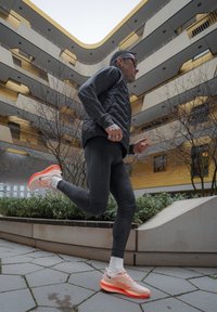 Veste matelassée noire, leggings noirs, et chaussures de course rose clair avec des accents orange. Courir dans une cour urbaine avec de la verdure.