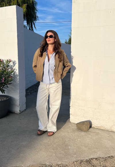 Chaqueta bomber marrón, camisa blanca de rayas y pantalones blancos holgados. Gafas de sol negras y sandalias marrones. Entorno al aire libre con paredes blancas y vegetación.