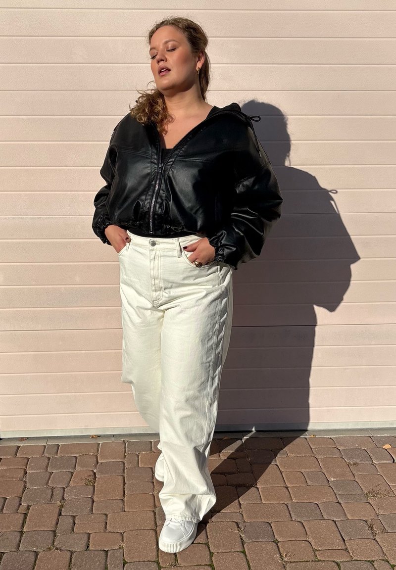 Zwarte leren cropped jacket, witte wijde jeans en witte sneakers. Zonlicht werpt een schaduw op een gestructureerd oppervlak.
