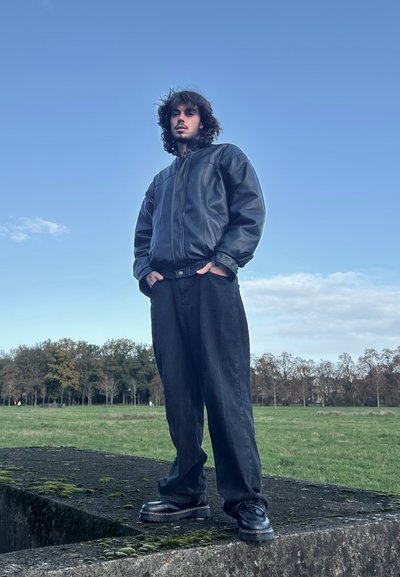 Chaqueta bomber de cuero negro, pantalones negros holgados y zapatos negros de suela gruesa. Entorno ventoso con vegetación y un cielo azul de fondo.
