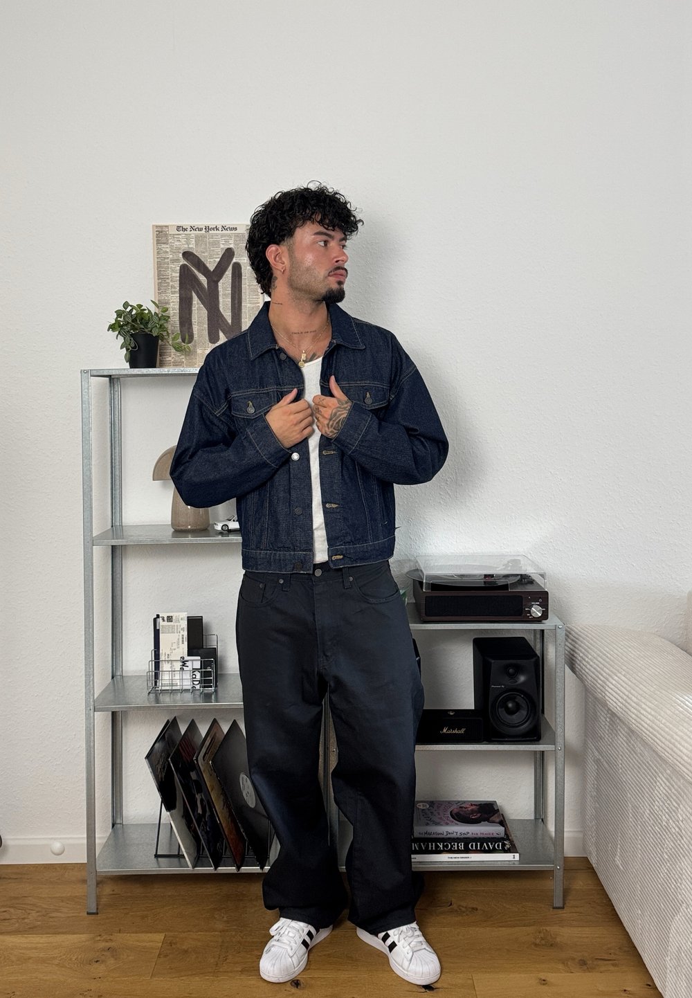 Jachetă din denim de culoare albastru închis, cu nasturi și două buzunare pe piept, purtată cu pantaloni largi negri și adidași albi. Stând într-o cameră.