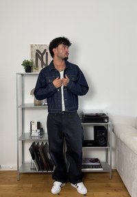 Jachetă din denim de culoare albastru închis, cu nasturi și două buzunare pe piept, purtată cu pantaloni largi negri și adidași albi. Stând într-o cameră.