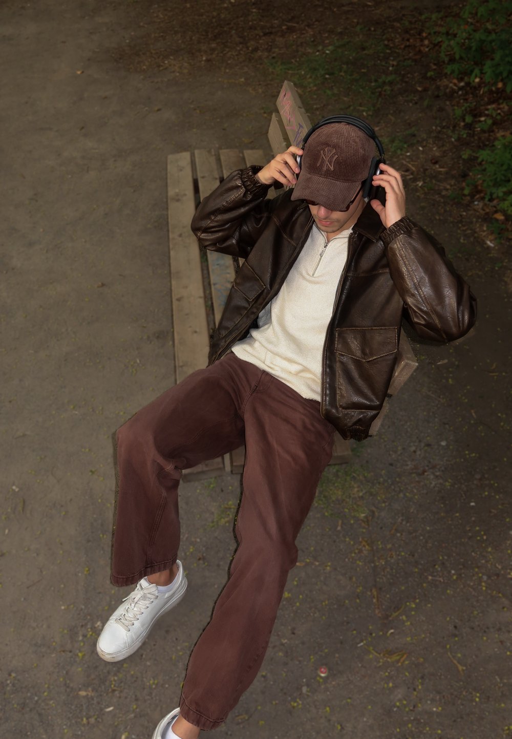 Brun læderjakke, beige zip-up skjorte, brune fløjlbukser, hvide sneakers og en brun kasket med høretelefoner, siddende på en træbænk.