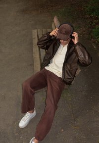 Brun læderjakke, beige zip-up skjorte, brune fløjlbukser, hvide sneakers og en brun kasket med høretelefoner, siddende på en træbænk.