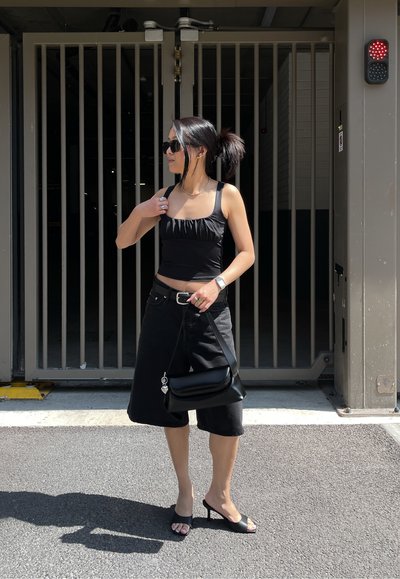 Top negro corto con detalles fruncidos, pantalones cortos negros hasta la rodilla, sandalias negras con tacón y un pequeño bolso negro. La luz del sol resalta la escena.