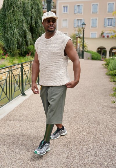 Top sin mangas de punto beige, pantalones cortos verde oliva y zapatillas grises con acentos verdes. Caminando por un sendero pavimentado cerca de vegetación.