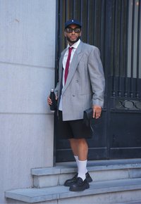 Γκρι διπλόμπροκο σακάκι, λευκό πουκάμισο, κόκκινη γραβάτα, μαύρες βερμούδες, λευκές κάλτσες, μαύρα παπούτσια, μαύρο καπέλο, κρατώντας ένα μαύρο μπουκάλι νερού και μία τσάντα.