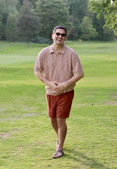 Camisa polo de manga corta, texturada, de color marrón claro con cuello; pantalones cortos naranjas; usando gafas de sol oscuras; caminando sobre un terreno cubierto de hierba.