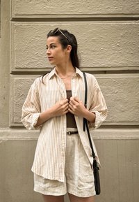 Light beige and cream striped shirt with rolled sleeves, paired with light linen shorts and a small black crossbody bag.