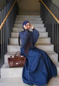 Jupe longue en jean avec un pull bleu texturé et un foulard. Sac en cuir marron à côté. Escalier en arrière-plan.