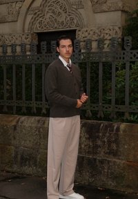 Brauner Cardigan über einem weißen Hemd, dunkler Krawatte, beigen Hosen und weißen Schuhen; stehend gegen eine Steinmauer mit grüner Umgebung im Hintergrund.