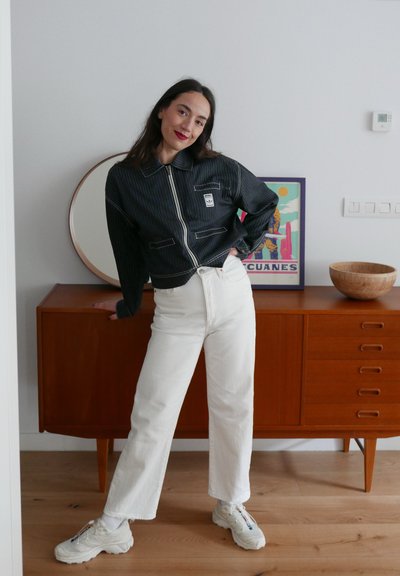 Chaqueta recortada a rayas blancas y negras con cremallera, combinada con pantalones blancos de cintura alta y zapatillas gruesas, ambientada en un interior moderno.