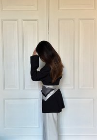 Sort blazer med lange ærmer, parret med hvide bukser med vidde. Et sølv- og sort mønstret tørklæde er bundet om taljen. Baggrunden er ensfarvet hvid.