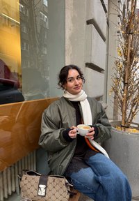 Grøn bomberjakke, sort sweater, cremefarvet tørklæde og blå jeans. Holder en hvid kop med kaffe. Brun mønstret håndtaske på gulvet.