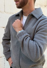 Άνδρας που φοράει γκρι φλις μπουφάν με φερμουάρ, άσπρο πουκάμισο από κάτω και ασημένιο ρολόι χειρός, στέκεται μπροστά σε έναν ανοιχτόχρωμο τοίχο από τούβλα.