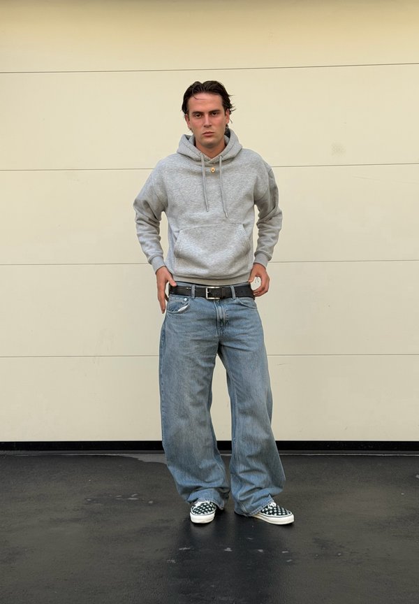 Joven de pie junto a una pared beige, vestido con sudadera gris con capucha, vaqueros azules holgados y zapatos tipo slip-on a cuadros en blanco y negro.