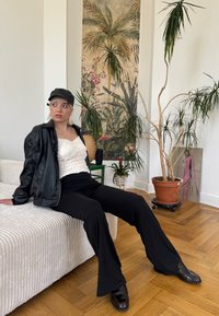 Pantaloni negri evazați, bluză albă texturată și o jachetă din piele neagră. Pantofi negri lucioși și o șapcă completează ținuta. Podea din lemn deschis și plante.