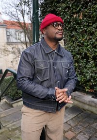 Άνδρας με κόκκινο σκουφάκι, γυαλιά και σκούρο τζιν μπουφάν στέκεται έξω δίπλα σε σκάλες και σε πράσινο τοίχο καλυμμένο με κισσό, με τα χέρια του σταυρωμένα μπροστά.