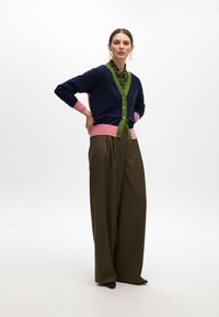 Il cardigan multicolore presenta sezioni blu navy, rosa e verdi con bottoni, indossato sopra una camicia verde con pattern e un pantalone largo color oliva.