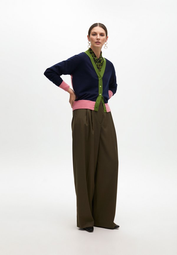 Il cardigan multicolore presenta sezioni blu navy, rosa e verdi con bottoni, indossato sopra una camicia verde con pattern e un pantalone largo color oliva.