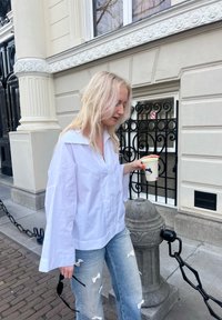 Camisa blanca de gran tamaño con mangas anchas, jeans azul claro con estampado de lazos, sosteniendo una taza, vista recortada de una calle con un poste ornamental.