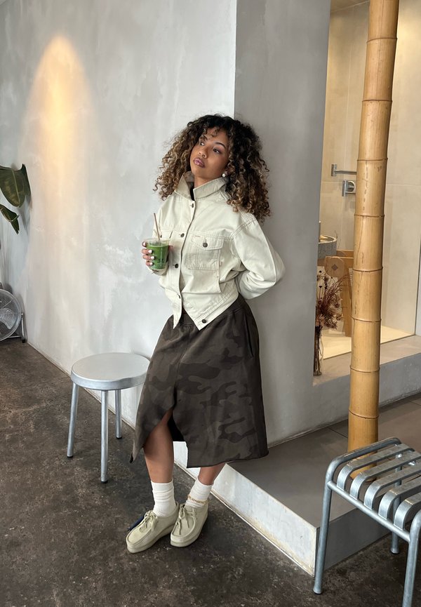 Mujer con cabello rizado se apoya en la pared sosteniendo una bebida verde, vistiendo una chaqueta clara, falda oscura, calcetines blancos y zapatos beige en un espacio interior minimalista.