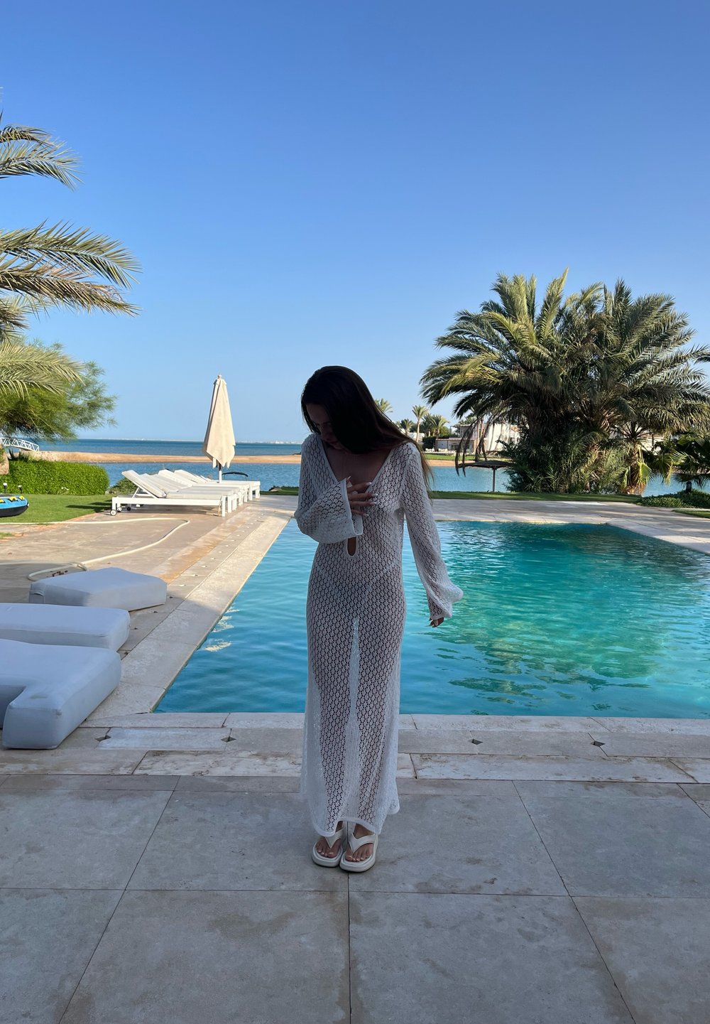 Abito maxi bianco, trasparente, con maniche lunghe, presenta un motivo a crochet e un dettaglio a occhiello, abbinato a infradito bianche, vicino a una piscina.