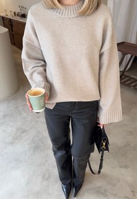 Άτομο που φοράει μπεζ πλεκτό πουλόβερ και μαύρο δερμάτινο παντελόνι, κρατώντας μια κούπα καφέ στο ένα χέρι και μια μαύρη τσάντα στο άλλο.