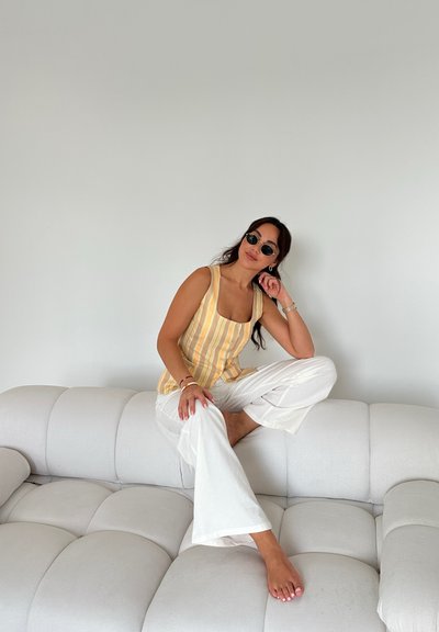 Top sin mangas a rayas amarillas y blancas con botones en la parte frontal, combinado con pantalones blancos holgados. Sentado en un sofá de terciopelo gris claro.