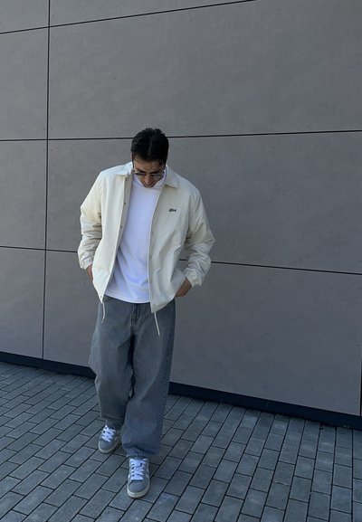 Hombre con chaqueta blanca, camisa blanca, vaqueros grises de pierna ancha y zapatillas grises caminando sobre suelo pavimentado cerca de una pared gris, manos en los bolsillos.