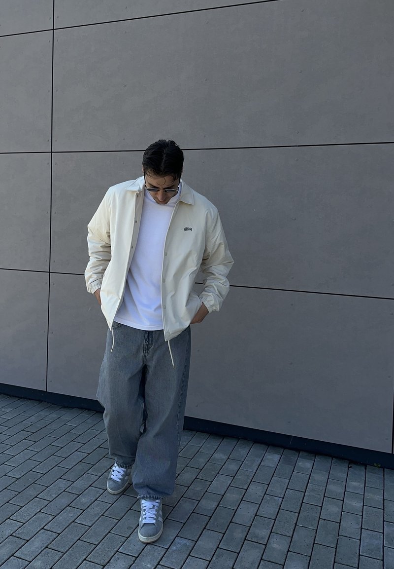 Homme portant une veste blanche, une chemise blanche, un jean gris à jambes larges et des baskets grises marchant sur un sol pavé près d'un mur gris, les mains dans les poches.