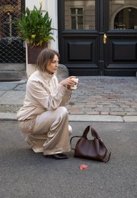 Giacca beige con zip e pantaloni abbinati, larghi, con bordo in pizzo. Borsa marrone a terra, che tiene una tazza di caffè. Strada in ciottoli.