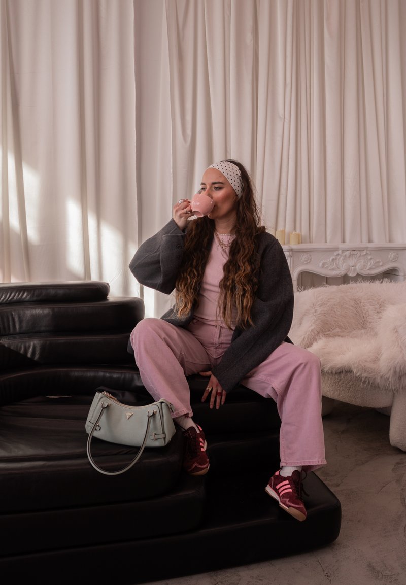 Jeune femme aux longs cheveux et bandeau, assise sur des marches en cuir noir, buvant dans une tasse rose, fauteuil moelleux et bougies derrière elle.