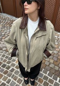 Beige bunda s hnědými koženými límci a manžetami, s kapsou na zip a uvolněným střihem. Nositá přes bílý strukturovaný top a černé kalhoty.